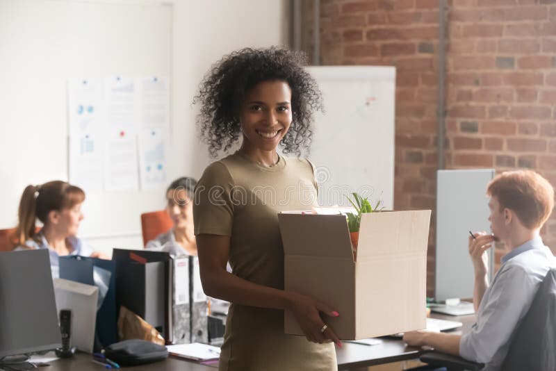 Smiling african American female newcomer holding box with personal belongings posing on first work day, happy excited black women newbie or intern take picture ready to unpack settle at new workplace. Smiling african American female newcomer holding box with personal belongings posing on first work day, happy excited black women newbie or intern take picture ready to unpack settle at new workplace