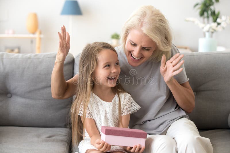 Excited grandchild happy to receive birthday present from loving old grandmother, smiling little kid holding gift box enjoys senior grandma surprise sitting on couch, granny congratulating child girl. Excited grandchild happy to receive birthday present from loving old grandmother, smiling little kid holding gift box enjoys senior grandma surprise sitting on couch, granny congratulating child girl