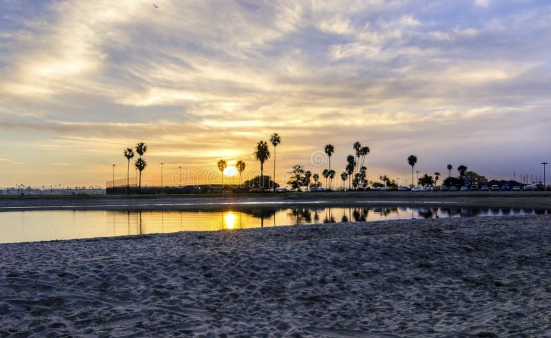The sunrise over Sail bay in Mission Bay over the Pacific beach in San Diego, California in the United States of America. A view of the sandy beach, palm trees and beautiful saltwater bay at sunset. The sunrise over Sail bay in Mission Bay over the Pacific beach in San Diego, California in the United States of America. A view of the sandy beach, palm trees and beautiful saltwater bay at sunset.