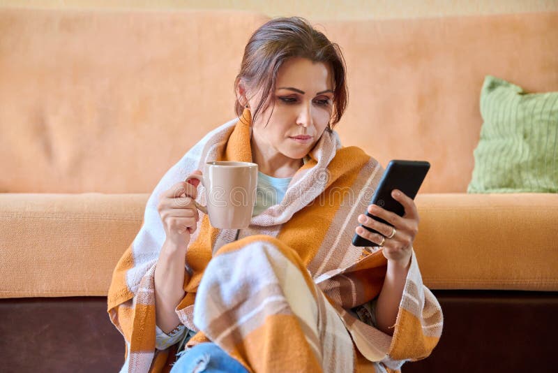 Autumn winter portrait of middle aged woman under warm blanket with cup and smartphone