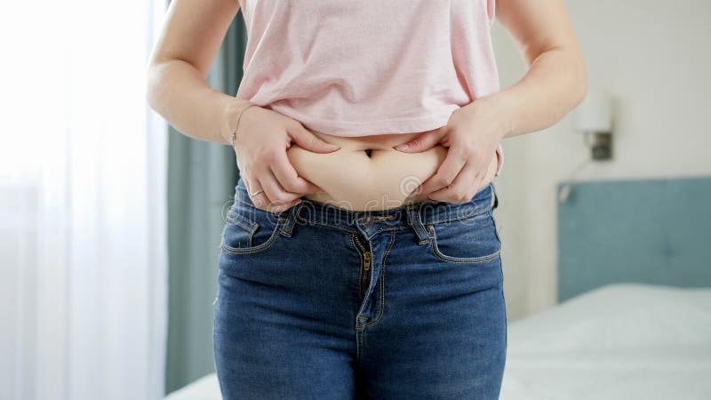 Avskärning av unga kvinnor på jeans som håller fettveck på sin buk. Begreppet övervikt, feta kostvanor hos hondjur och royaltyfria foton