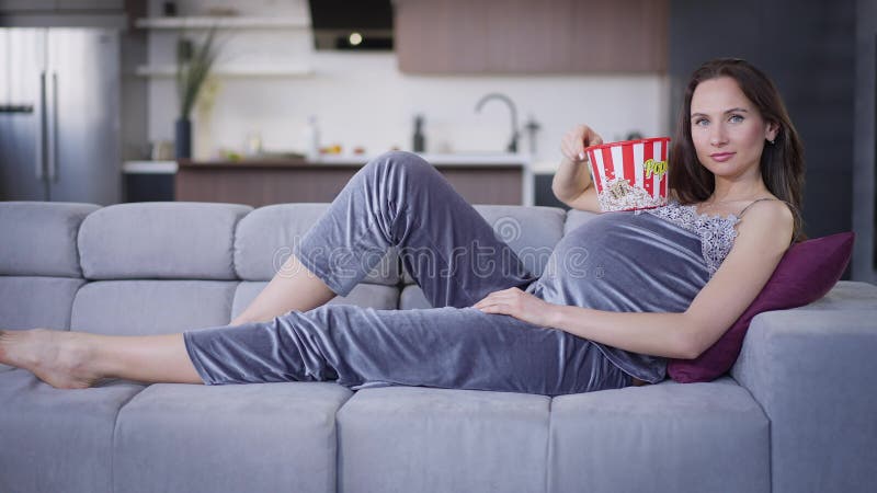 Avslaxad gravid kaukasisk ung kvinna som ligger på bekvämt soffa med popcornhink och tittar på kameran. vid skott