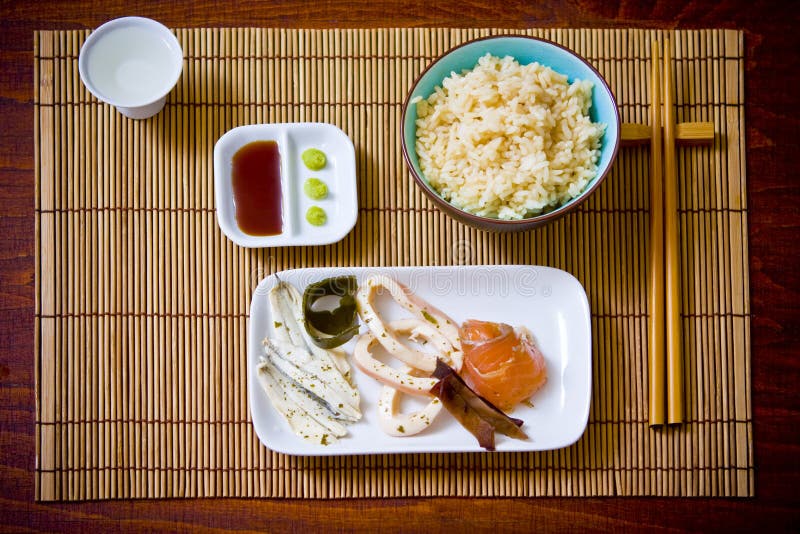 Asian food including rice, fish, wasabi and sake with chopsticks on a bamboo place mat. Asian food including rice, fish, wasabi and sake with chopsticks on a bamboo place mat.