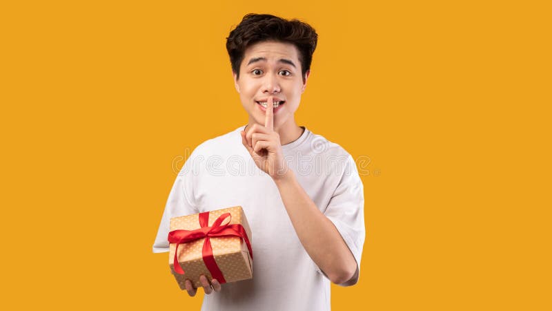 Surprise Gift. Portrait of smiling young asian guy holding wrapped present box and showing silence sign, isolated over orange yellow studio background, greeting and celebrating special day or event. Surprise Gift. Portrait of smiling young asian guy holding wrapped present box and showing silence sign, isolated over orange yellow studio background, greeting and celebrating special day or event