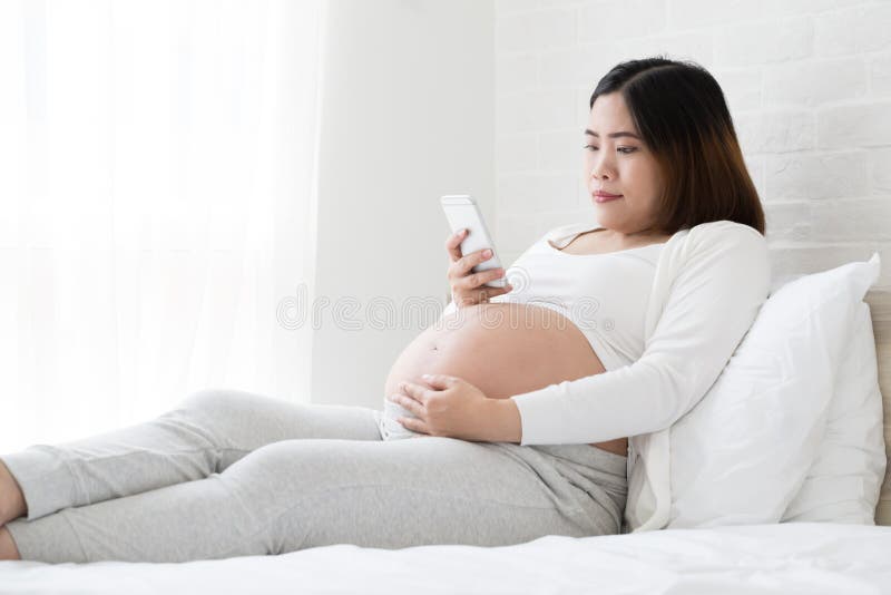 Aziatische zwangere vrouw met smartphone en thuis het houden op het bed bijgewerkt, Gebruikend mobiele telefoon tijdens zwangersc stock fotografie Aziatische zwangere vrouw met smartphone en thuis het houden op het bed bijgewerkt, Gebruikend mobiele telefoon tijdens zwangersc stock fotografie