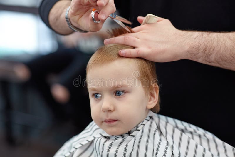 Serious Cute Blond Baby Boy With Blue Eyes In A Barber Shop Having Haircut By Hairdresser Hands Of Stylist With Tools Scissor B Image Stock Image Du Client Beaute