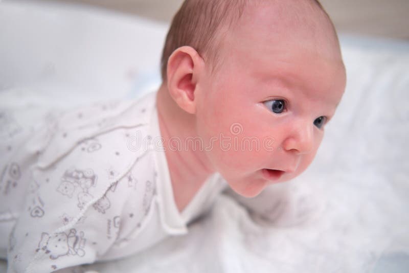 A baby aged 1 month learns to keep his head lying on his stomach. Caucasian boy child in a home white bedroom