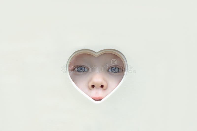 The baby face, eyes and nose peep out of a heart-shaped hole in the center. A child looks into the camera through a stock image The baby face, eyes and nose peep out of a heart-shaped hole in the center. A child looks into the camera through a stock image