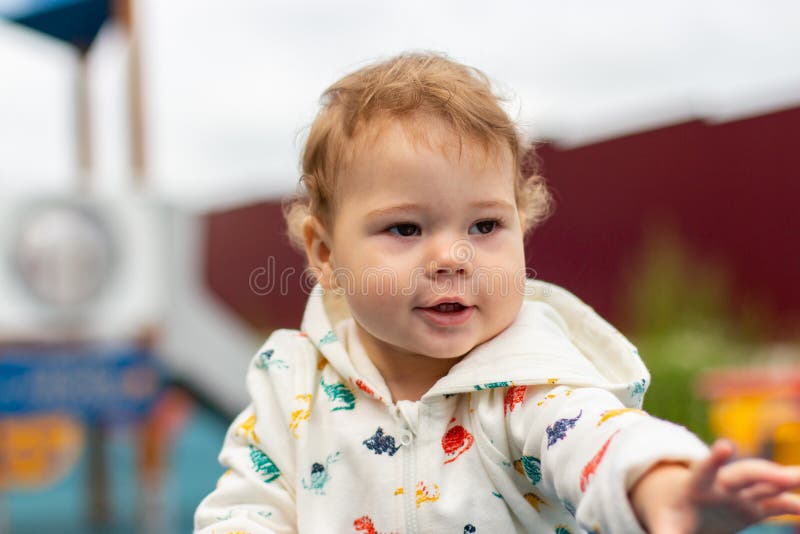 Baby girl in een wit jasje op de speelplaats buiten Gefeliciteerd, blanke baby 1 jaar, horizontaal portret Krulharen royalty-vrije stock fotografie Baby girl in een wit jasje op de speelplaats buiten Gefeliciteerd, blanke baby 1 jaar, horizontaal portret Krulharen royalty-vrije stock fotografie