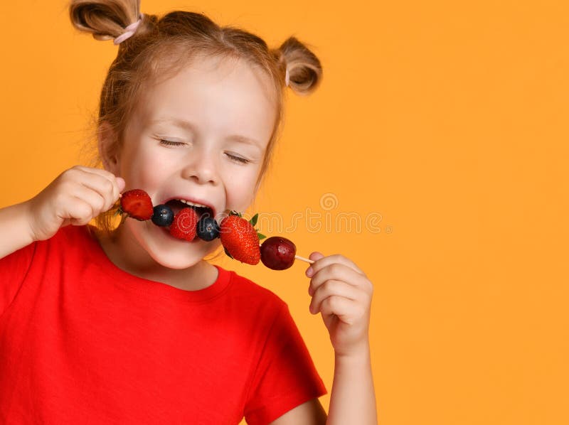 Baby girl kid in red t-shirt holding smelling biting eating fresh berries dessert with cherry strawberry raspberry on skewer stock image Baby girl kid in red t-shirt holding smelling biting eating fresh berries dessert with cherry strawberry raspberry on skewer stock image