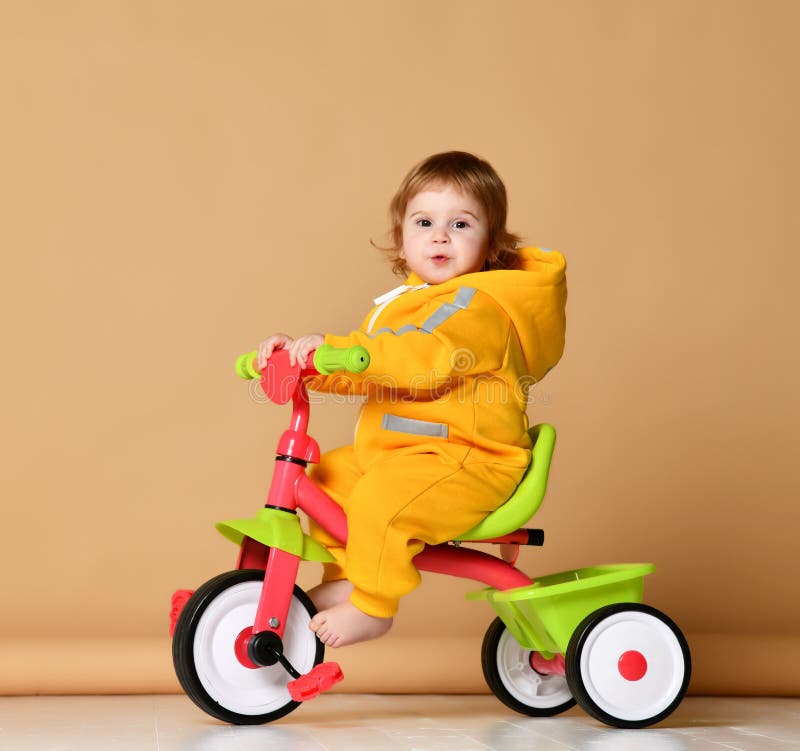 Baby girl kid riding her first bicycle tricycle in warm yellow overalls looking up on light grey background