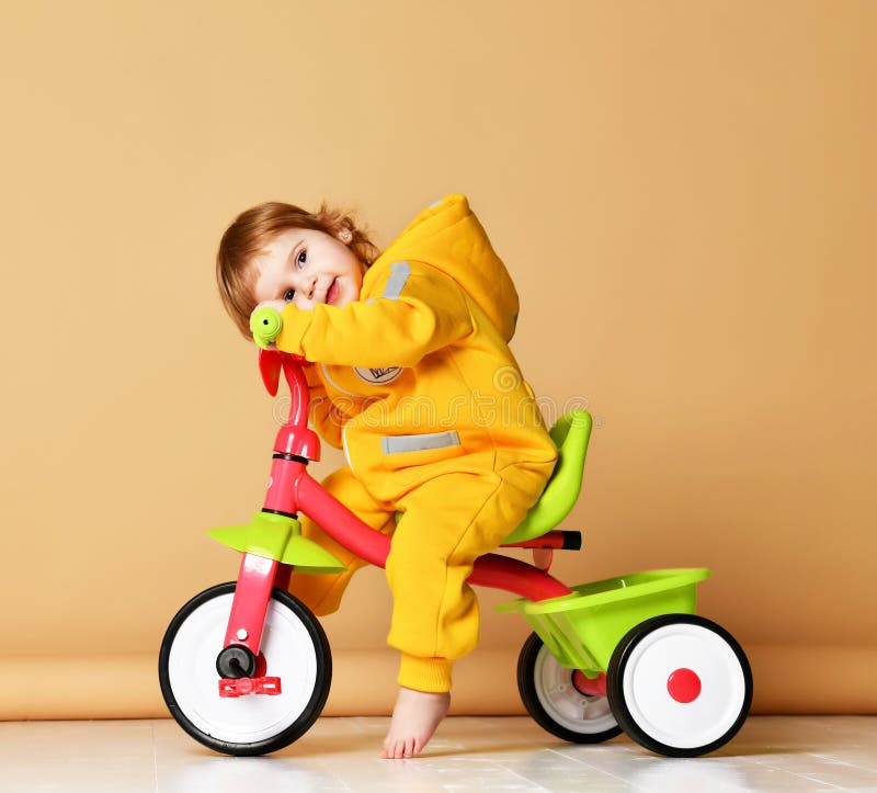 Baby girl kid toddler riding her first bicycle tricycle in warm yellow overalls looking up on light grey background. Baby girl kid toddler riding her first bicycle tricycle in warm yellow overalls looking up on light grey background