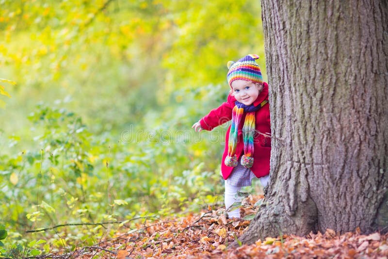 Sweet funny baby girl in a red coat and colorful hat and knitted scarf hiding behind a big old tree in a beautiful autumn park with yellow leaves. Sweet funny baby girl in a red coat and colorful hat and knitted scarf hiding behind a big old tree in a beautiful autumn park with yellow leaves