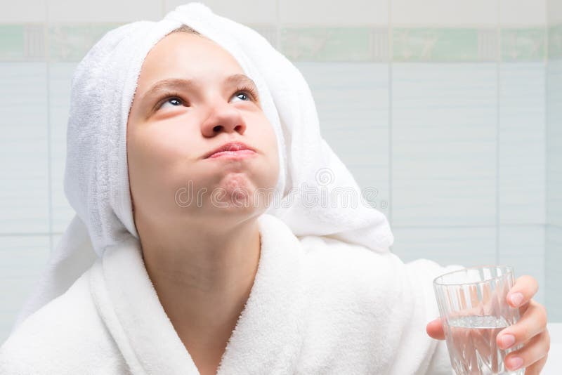 Baby girl, in a white robe and towel on her head, in the bathroom, holding a glass of water and rinsing her mouth, after cleaning