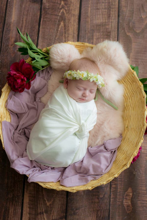 Baby girl in a wicker basket of vine decorated with burgundy peonies in a light winding and a flower wreath on her head.