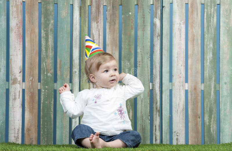 Baby with carnival hat seated in the garden, against a fence. Baby with carnival hat seated in the garden, against a fence