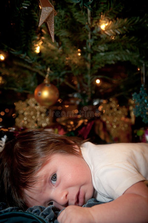A baby with a scared expression laying down under a Christmas tree. The image has a vertical orientation and copy space. A baby with a scared expression laying down under a Christmas tree. The image has a vertical orientation and copy space.