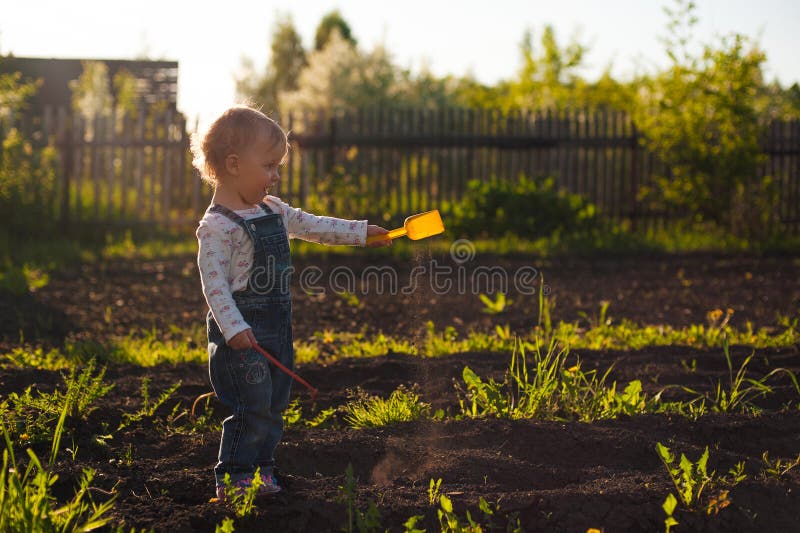 Baby sitting on the ground with shovel and rake in sunlight. Cute summer blond girl in the garden royalty free stock image Baby sitting on the ground with shovel and rake in sunlight. Cute summer blond girl in the garden royalty free stock image