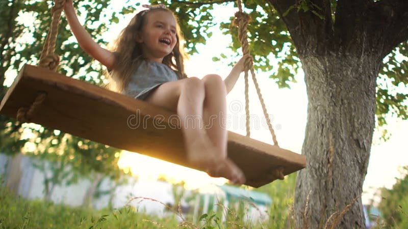 The baby swings on a swing in the summer against the backdrop of the setting sun. Someone tries to grab her by the legs