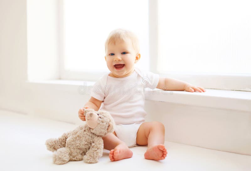 Baby with teddy bear toy sitting home in white room near wind