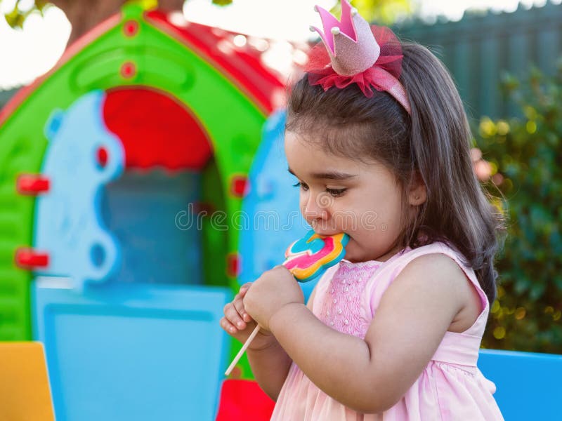 Baby toddler girl eating a large colorful lollipop dressed in pink dress as princess or queen