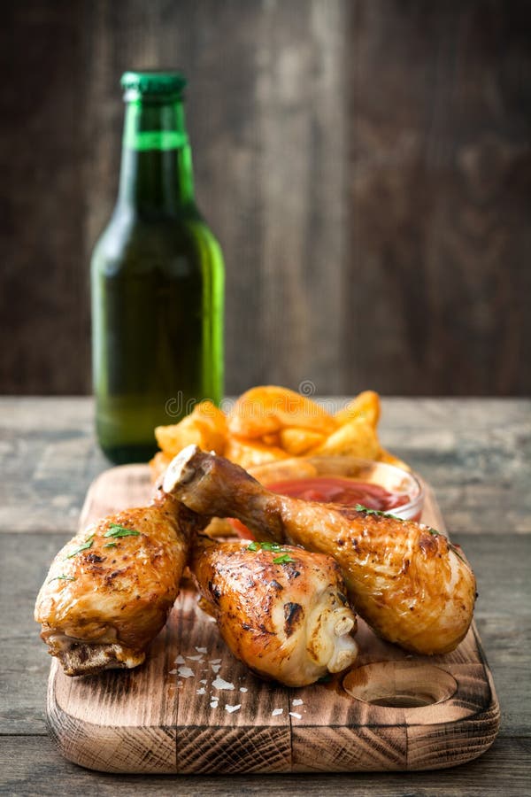 Bacchette e patatine fritte di pollo arrosto deliziose, birra sul tagliere e fondo di legno del fondo della tavola fotografia stock libera da diritti Bacchette e patatine fritte di pollo arrosto deliziose, birra sul tagliere e fondo di legno del fondo della tavola fotografia stock libera da diritti