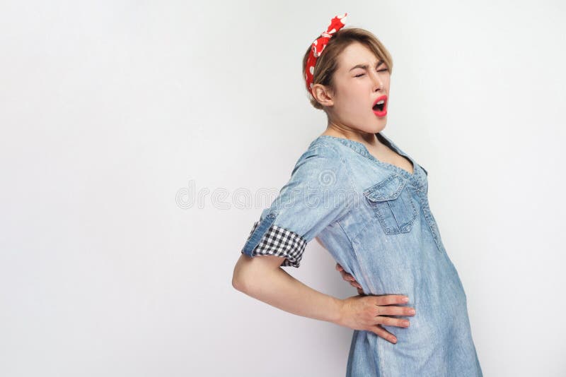 Back or kidney pain. profile side view portrait of young woman in casual blue denim shirt with red headband standing and holding her painful back. indoor studio shot, isolated on white background