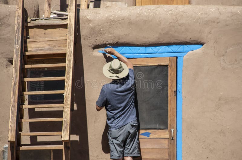 Back of Native American man in shorts and tee-shirt and sun hat painting bright blue-turquoise around the door of a mud adobe