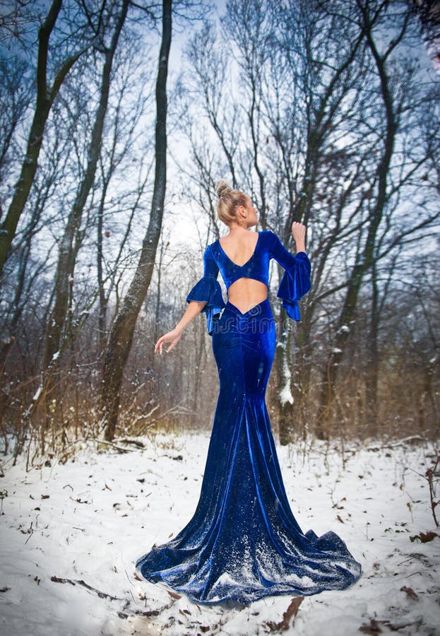 Back side view of lady in long blue dress posing in winter scenery, royal look. Fashionable blonde woman with forest in background, outdoor shot. Glamorous fair hair female in nature - princess style