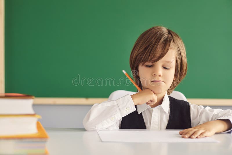 Serious little boy writing something in notebook at desk in classroom, copy space. Hardworking child studying at table. Back to school concept. Serious little boy writing something in notebook at desk in classroom, copy space. Hardworking child studying at table. Back to school concept