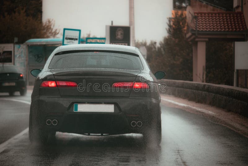 Back view of a black car on the road. Brake lights turned on under braking, cloudy dark day with heavy rain. Water puddles on the