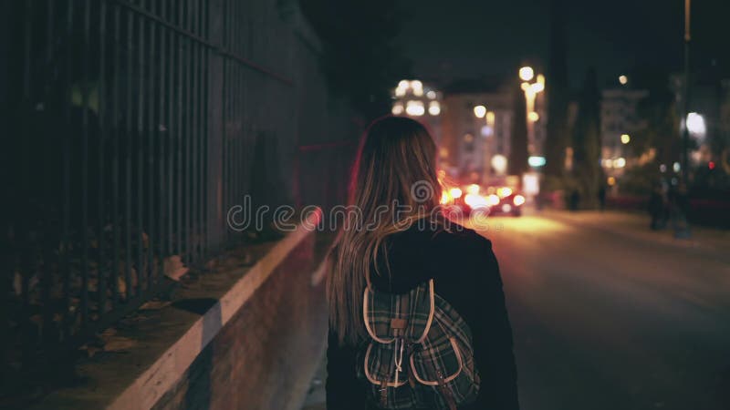 Back view of brunette woman walking near the road at the traffic time. Girl goes through the city late at night alone.