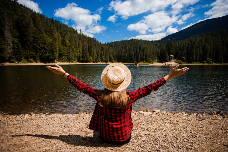 Back view of female tourist in hat with hands up enjoying freedom and amazing scenery of mountain lake.