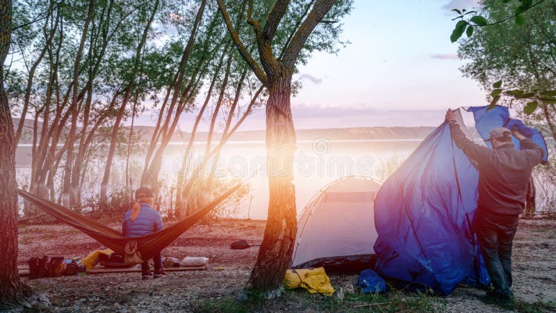 Back view of girl relaxing on olive hammock between two trees enjoying the view at the lake in summer evening a man sets up a tent royalty free stock photography Back view of girl relaxing on olive hammock between two trees enjoying the view at the lake in summer evening a man sets up a tent royalty free stock photography