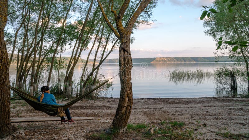 Back view of girl relaxing on olive hammock between two trees enjoying the view at the lake in summer morning. Copy space. Bakota royalty free stock photo Back view of girl relaxing on olive hammock between two trees enjoying the view at the lake in summer morning. Copy space. Bakota royalty free stock photo