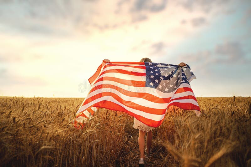 Back view of a girl in white dress wearing an American flag while running in a beautiful wheat field