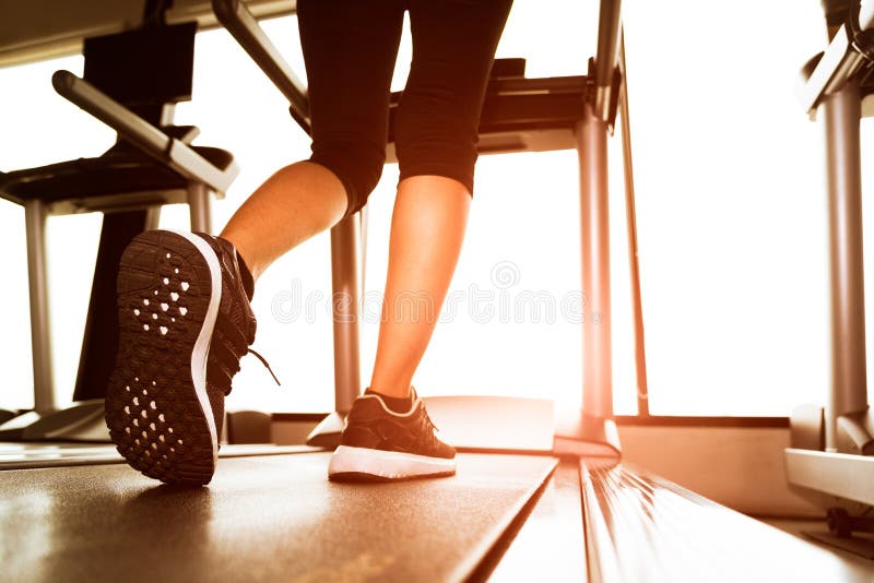 Back view of lower body legs of fitness girl running on machine or treadmill in fitness gym with sun ray. Warm tone. Healthy and Exercise activity concept. Workout and Strength training theme