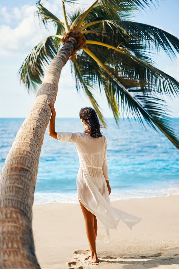 Back view of pretty young woman relax and enjoy sea standing under palm tree on tropical beach