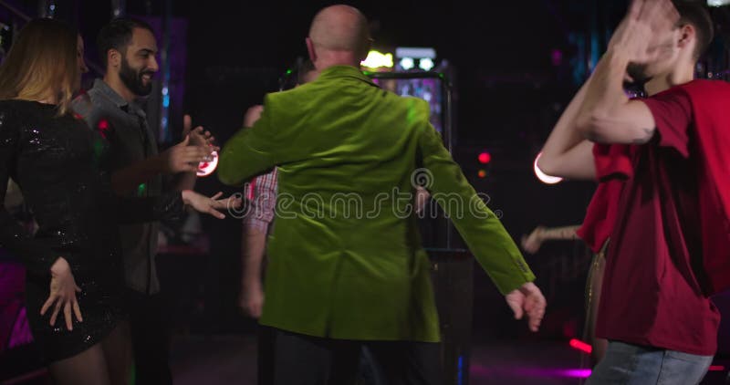 Back view of stylish mature Caucasian man in green jacket dancing in night club surrounded by young people. Cheerful