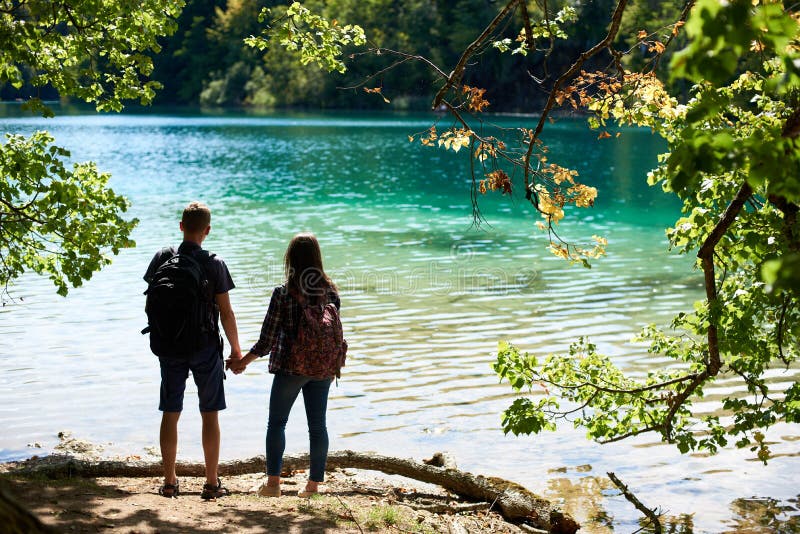 Back view of tourist couple boy and girl with backpacks standing on river bank stock photo Back view of tourist couple boy and girl with backpacks standing on river bank stock photo