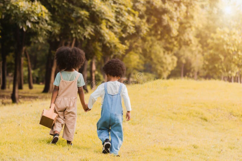 Back view of two adorable african children, girl and boy holding hand and walking together in garden