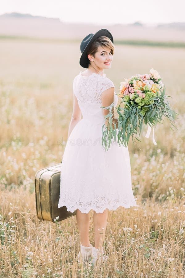 The back view of the vintage dressed bride holding the wedding bouquet, carrying the old-fashioned suitcase and smiling