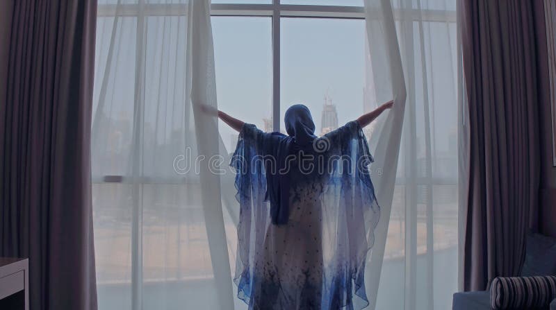 Back view of woman opening curtains and looking out of window to cityscape at hotel in the morning. Modern buildings at the Dubai city. High quality photo