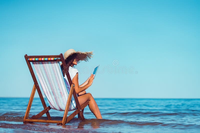 Back view of woman in sunhat who using laptop while lying on the beach chaise longue at the seaside royalty free stock image