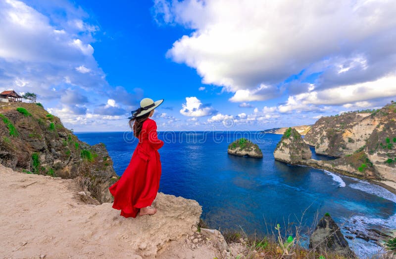Back view woman wearing a red dress on the edge of a cliff looking at Diamond beach, Nusa Penida. Scenic view of Diamond Beach in Nusa Penida Bali, Indonesia.