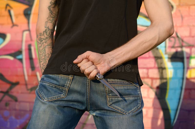 Back view of young caucasian man with knife in his hand against ghetto brick wall with graffiti paintings. Concept of criminal