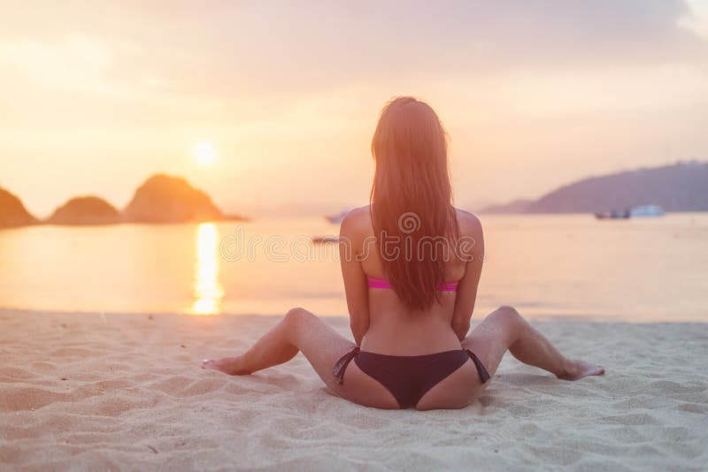 Back view of young female with long brown hair sitting on beach legs spread far apart in black bikini panties admiring