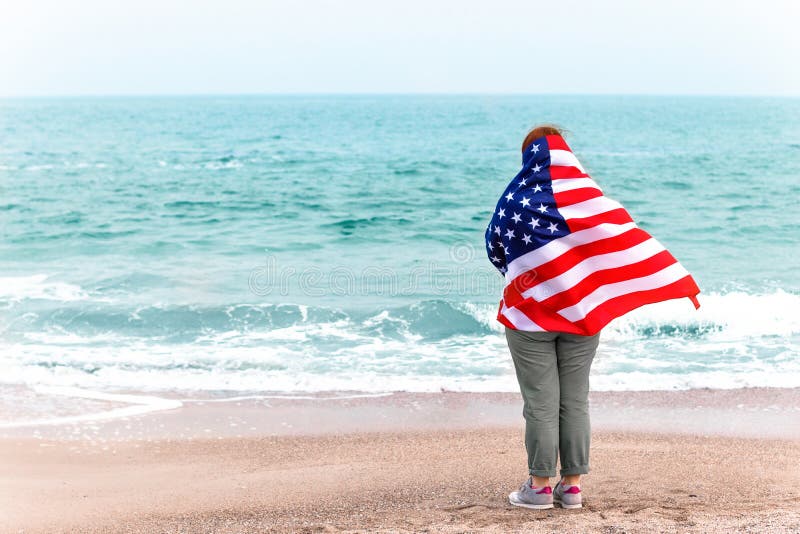 Back view of a young girl with an American flag looks at the sea. Back view of young girl with an American flag looks at the sea. America flag holiday concept stock photography Back view of a young girl with an American flag looks at the sea. Back view of young girl with an American flag looks at the sea. America flag holiday concept stock photography