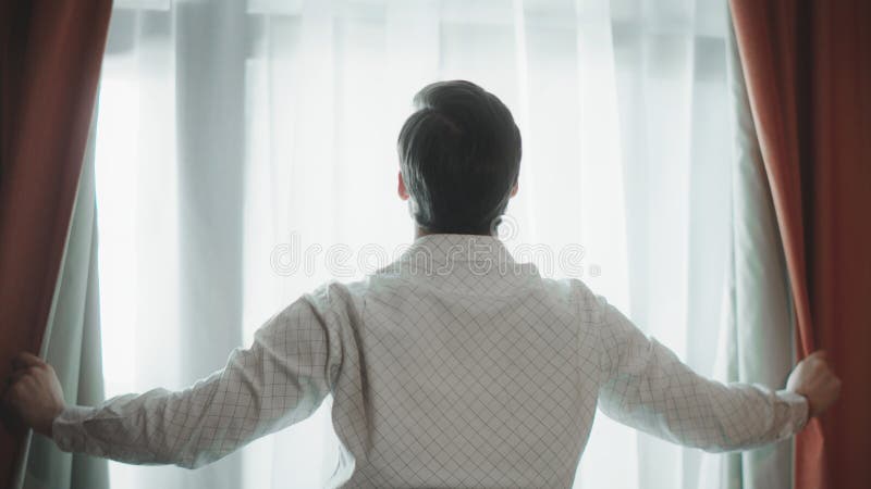 Back view of young man opening curtains at hotel room. Close up of handsome business man standing by window. Successful businessman opening curtains at modern apartment. Back view of young man opening curtains at hotel room. Close up of handsome business man standing by window. Successful businessman opening curtains at modern apartment