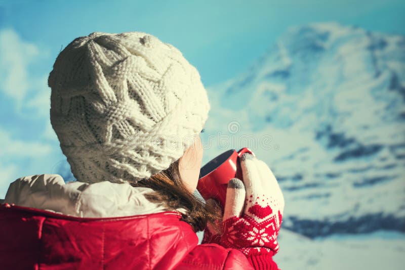 Back view of young woman in a winter knitted white cap, jacket and Christmas gloves drinking hot coffee or chocolate from  cup